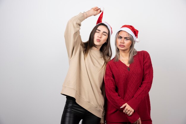 Imagen de una mujer feliz sosteniendo un gorro de Papá Noel cerca de su amiga triste mujer bonita.