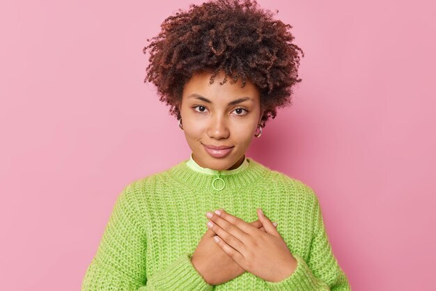 La imagen de una mujer guapa con el pelo rizado presiona las manos en el pecho hace que el gesto de gratitud se vea con aprecio tiene una cara agradecida viste un suéter de punto casual aislado sobre fondo rosa