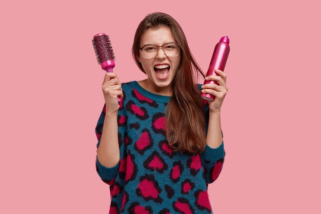 Imagen de mujer hermosa desesperada grita enojada, no puedo peinarse, está enojada con el peluquero, lleva peine y spray, tiene el pelo largo y liso