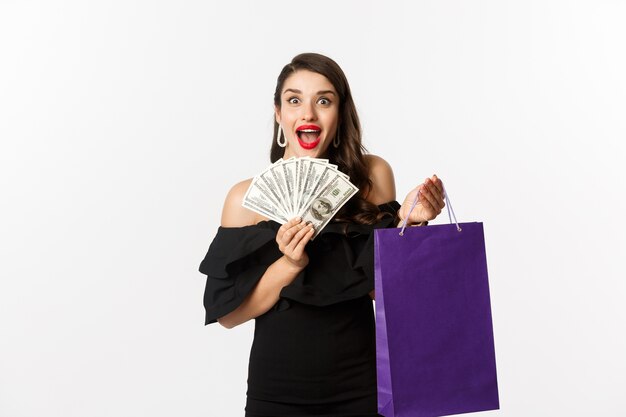 Imagen de mujer hermosa emocionada en vestido negro yendo de compras, sosteniendo el bolso y dólares, de pie sobre fondo blanco.