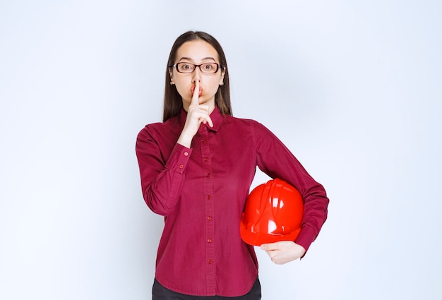 Imagen de mujer hermosa con gafas sosteniendo un casco y haciendo un signo de silencio.