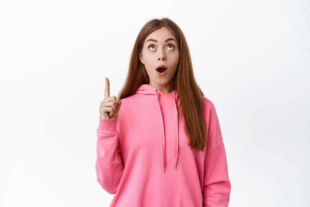 Imagen de una mujer joven asombrada mirando una oferta de promoción increíble, jadeando y apuntando con el dedo hacia arriba en el espacio de la copia en la parte superior, de pie sobre la pared blanca