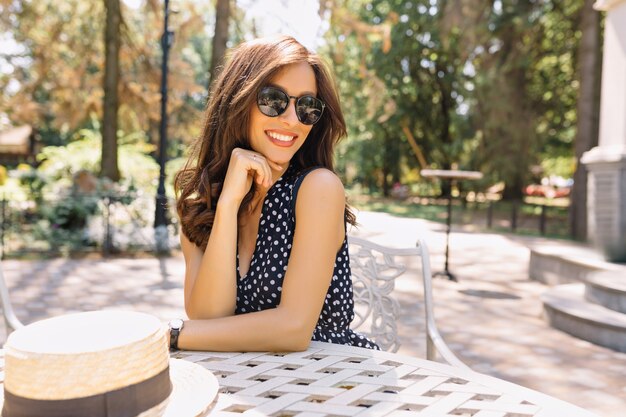 Imagen de una mujer joven con un cabello hermoso y una sonrisa encantadora está sentada en la cafetería de verano a la luz del sol. Lleva un bonito vestido de verano y gafas de sol negras.