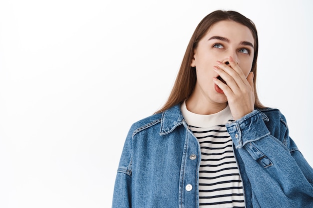 Imagen de una mujer joven cansada, una niña aburrida que bosteza y mira el texto promocional, siente fatiga, se despierta temprano en la mañana sin café, de pie sobre una pared blanca