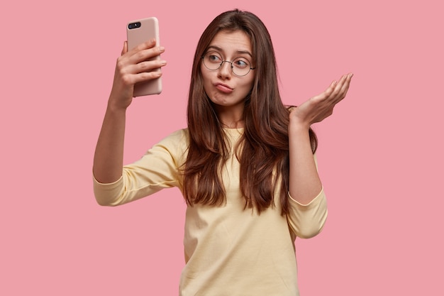 Imagen de mujer joven caucásica bastante vacilante mira con apatía de teléfono inteligente, toma selfie o hace videollamada