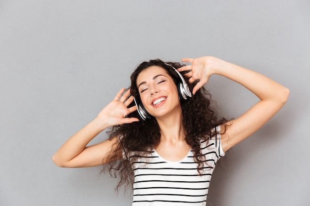 Imagen de una mujer joven complacida en camiseta a rayas que disfruta de sus canciones favoritas escuchando melodías a través de auriculares con los ojos cerrados sobre la pared gris