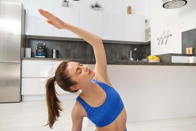 Imagen de una mujer joven concentrada haciendo ejercicios deportivos desde casa en sostén deportivo azul y piernas sentadas