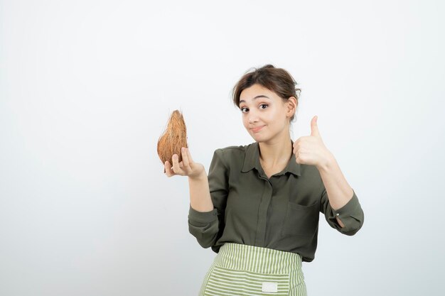 Imagen de mujer joven en delantal mostrando el pulgar hacia arriba y sosteniendo un coco. foto de alta calidad