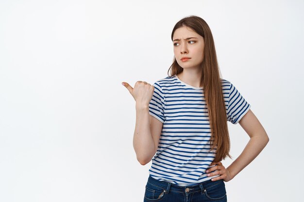 Imagen de una mujer joven escéptica con el ceño fruncido, señalando y mirando a la izquierda con expresión dudosa y poco impresionada, de pie en blanco.