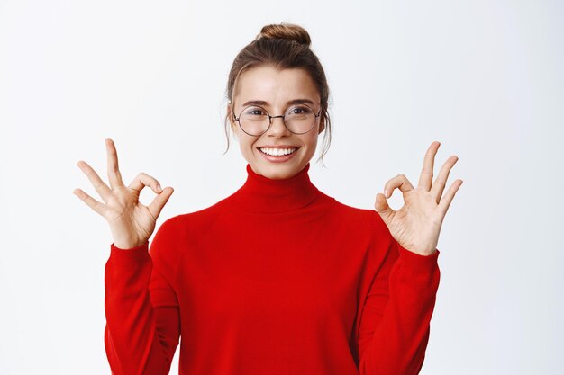 Imagen de una mujer joven guapa con gafas, sonriendo satisfecha, mostrando signos de aprobación en señal de aprobación, como algo, di bien, haciendo un gesto de sí en blanco