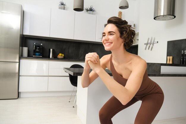 Imagen de una mujer joven haciendo sentadillas en casa haciendo ejercicio en una alfombra de goma en una habitación luminosa en el interior con ropa activa