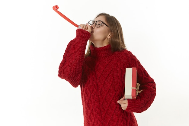 Imagen de una mujer joven hermosa alegre en estado de ánimo festivo celebrando el año nuevo o la Navidad, vistiendo un suéter de punto cálido y anteojos, sosteniendo una caja y soplando un cuerno de fiesta de papel, divirtiéndose