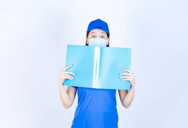 Imagen de mujer joven en máscara médica posando con carpeta azul sobre pared blanca.