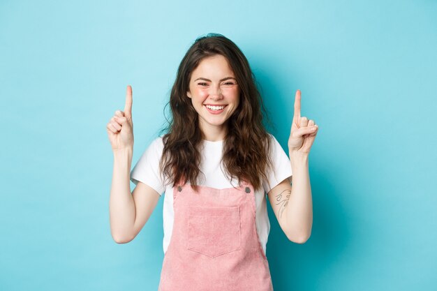 Imagen de mujer joven sonriente feliz con pelo rizado, riendo y señalando con el dedo hacia el banner del logo, mostrando trato promocional, de pie contra el fondo azul.