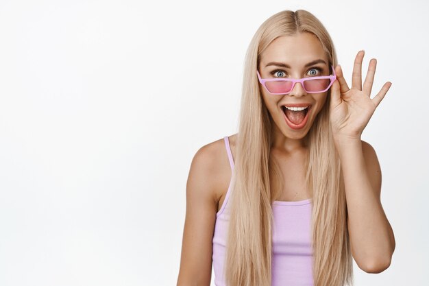 Imagen de una mujer joven sorprendida y feliz mirando con sus gafas de sol rosas, sonriendo y mirando emocionado a la venta super cool en blanco