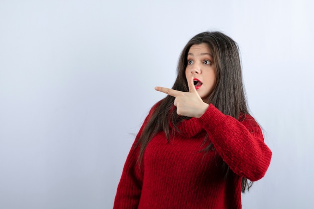 Imagen de una mujer joven sorprendida en suéter rojo apuntando hacia afuera