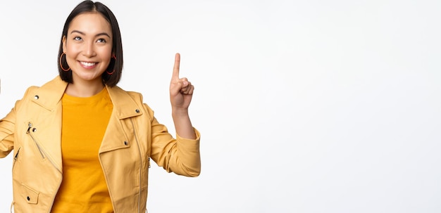 Imagen de una mujer morena asiática sonriente señalando con el dedo hacia arriba mostrando un anuncio con cara feliz posando sobre fondo blanco