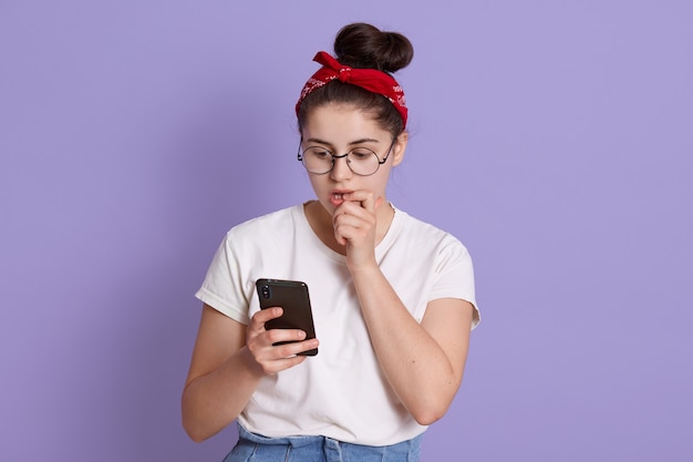 Imagen de mujer morena asustada con nudo y diadema mordiendo su dedo, siente miedo, sosteniendo un teléfono inteligente, viste camiseta blanca