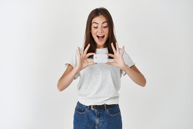 Imagen de una mujer morena feliz mostrando una tarjeta de crédito de plástico y mirándola con alegría y asombro de pie en camiseta contra fondo blanco