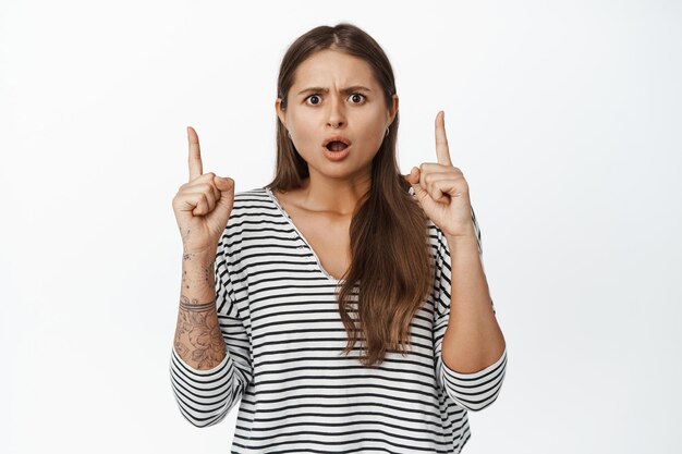 La imagen de una mujer muestra frustración mientras señala con los dedos hacia arriba, frunce las cejas y se ve conmocionado, fondo blanco.