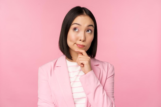 Imagen de una mujer de negocios asiática parada en una pose de pensamiento haciendo una lluvia de ideas con traje Empresaria vendedora coreana posando sobre un fondo rosa