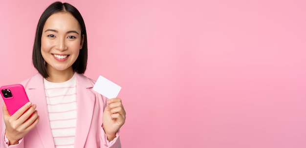 Imagen de una mujer de negocios asiática sonriente y feliz que muestra el pago con tarjeta de crédito en línea en un pedido de aplicación de teléfono inteligente con un teléfono móvil de pie contra un fondo rosa
