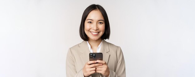 Imagen de una mujer de negocios asiática con traje sosteniendo un teléfono móvil usando una aplicación de teléfono inteligente sonriendo a la cámara de fondo blanco