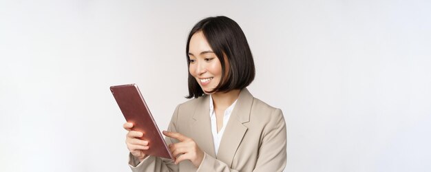 Imagen de una mujer de negocios asiática usando una tableta digital mirando un aparato y sonriendo trabajando de pie contra un fondo blanco