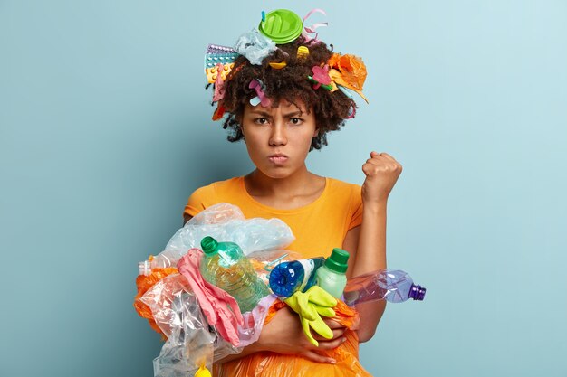 Imagen de una mujer negra molesta que levanta el puño cerrado, exige ser respetuoso con el medio ambiente, tiene una expresión facial gruñona, lleva desechos plásticos, usa objetos para reciclar, se para sobre una pared azul