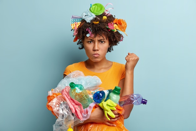 Imagen de una mujer negra molesta que levanta el puño cerrado, exige ser respetuoso con el medio ambiente, tiene una expresión facial gruñona, lleva desechos plásticos, usa objetos para reciclar, se para sobre una pared azul