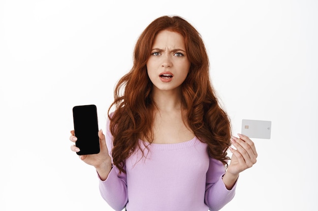 Imagen de una mujer pelirroja frustrada frunciendo el ceño, mostrando la pantalla vacía del teléfono inteligente y la tarjeta de crédito, mirando decepcionada, quejándose, parada sobre fondo blanco