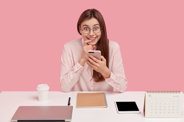 Imagen de mujer positiva con expresión alegre, mantiene el dedo cerca de los labios, sostiene celular, toma café, mensajes en redes sociales