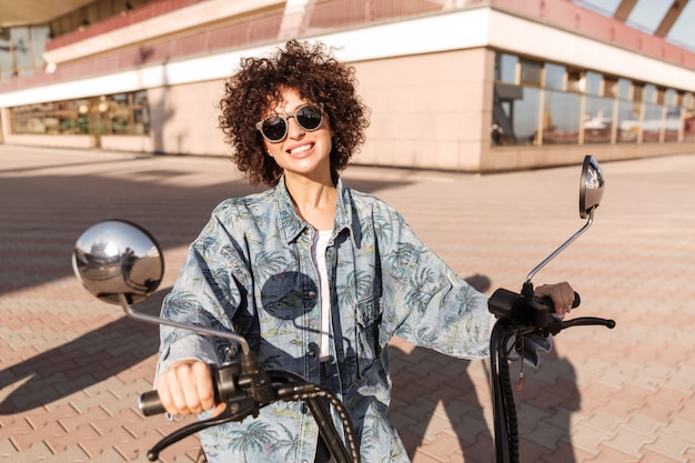 Imagen de mujer rizada alegre en gafas de sol sentado en moto