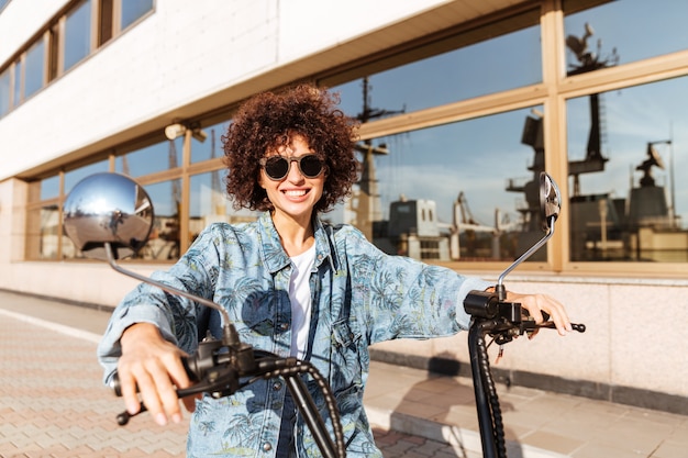 Imagen de mujer rizada feliz en gafas de sol sentado en moto moderna al aire libre