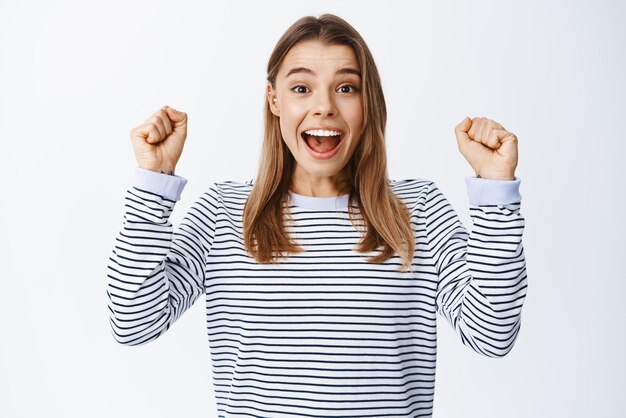 Imagen de una mujer rubia emocionada ganando y celebrando el logro levantando las manos y gritando de alegría lograr el objetivo de pie sobre fondo blanco