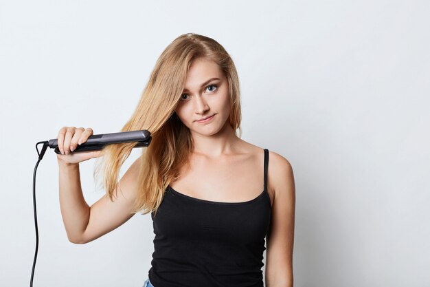 La imagen de una mujer rubia vestida de negro usa una plancha para el cabello, trata de peinarse antes de ir a la fiesta. La mujer hace peinado en casa, usa plancha para el cabello. Vendedor femenino anuncia productos de belleza