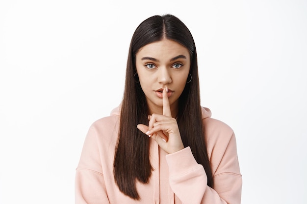 Imagen de una mujer seria que hace un gesto tabú muestra el signo shh shush en los labios y mira fijamente a la cámara pidiendo una posición secreta con capucha contra fondo blanco