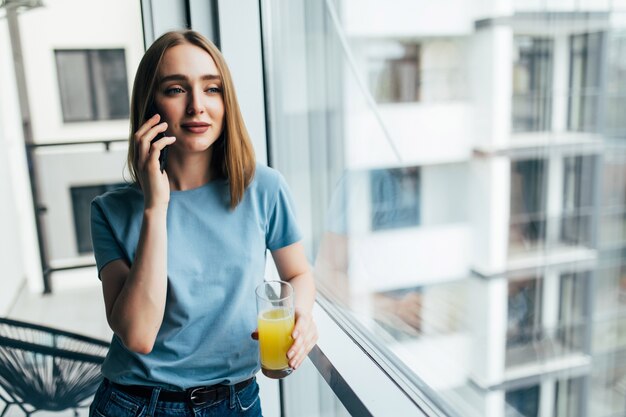 Imagen de mujer sonriente hablando por teléfono celular y bebiendo jugo mientras está de pie cerca de la ventana en el interior