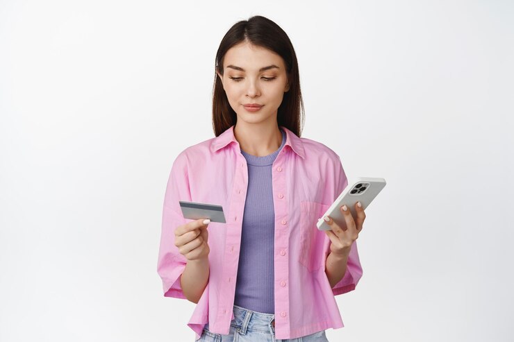 Foto gratuito imagen de una mujer sonriente haciendo una compra en un teléfono móvil mirando la tarjeta de crédito mientras pide algo en una aplicación de teléfono inteligente de pie sobre fondo blanco