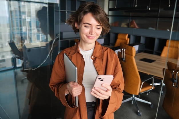 Foto gratuita imagen de una mujer trabajadora elegante revisando sus mensajes en un teléfono inteligente sosteniendo una computadora portátil parada en un