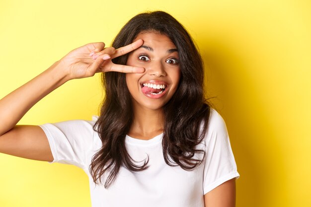 Imagen de una niña afroamericana adolescente tonta y linda, mostrando el signo de la paz y sonriendo, de pie sobre fondo amarillo