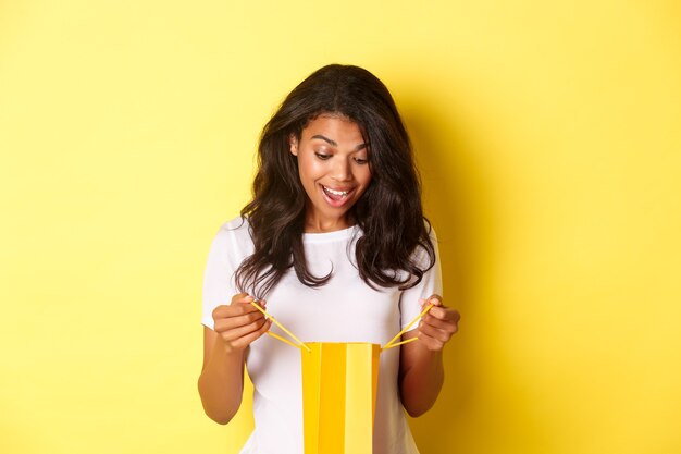 Imagen de niña afroamericana feliz y sorprendida, recibiendo un regalo, mirando dentro de compras mal con asombro, de pie sobre fondo amarillo
