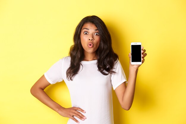 Imagen de una niña afroamericana sorprendida mirando fascinada y mostrando la pantalla del teléfono inteligente de pie