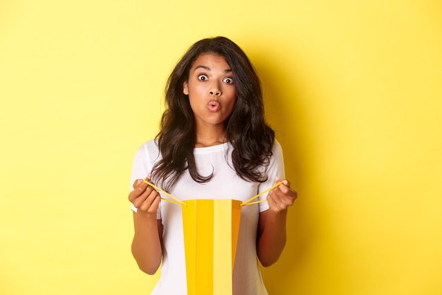 Imagen de niña afroamericana sorprendida recibir un regalo de vacaciones, abrir una bolsa de compras y mirar asombrada, de pie sobre fondo amarillo.