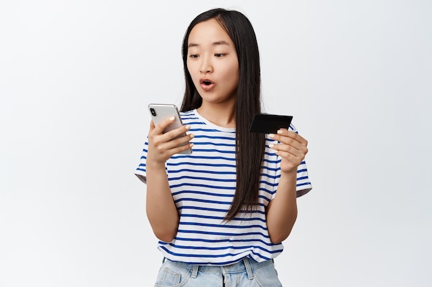 Imagen de niña asiática con tarjeta de crédito, se ve emocionada en la pantalla del teléfono móvil, pagando por compras en línea, usando la aplicación de la tienda, de pie en blanco.