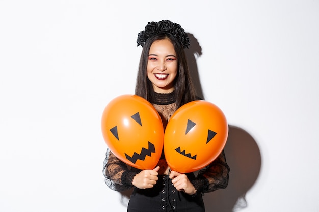 Imagen de niña asiática en traje de bruja malvada sosteniendo dos globos naranjas con caras aterradoras, celebrando halloween, de pie sobre fondo blanco.