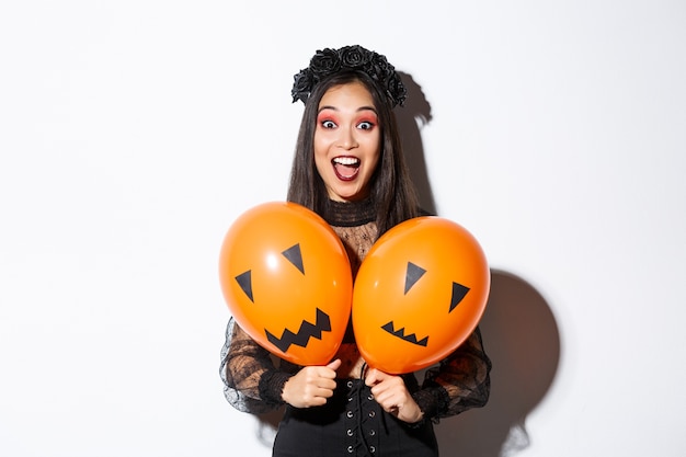 Foto gratuita imagen de niña asiática en traje de bruja malvada sosteniendo dos globos naranjas con caras aterradoras, celebrando halloween, de pie sobre fondo blanco.