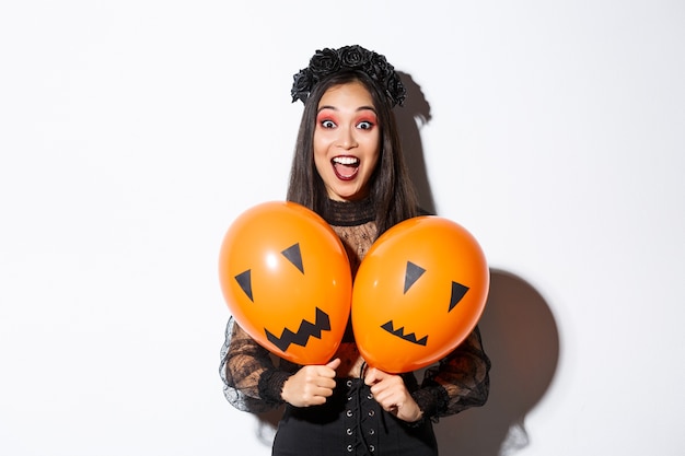 Imagen de niña asiática en traje de bruja malvada sosteniendo dos globos naranjas con caras aterradoras, celebrando halloween, de pie sobre fondo blanco.