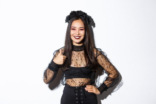 Imagen de niña bonita asiática satisfecha en traje de halloween mostrando pulgar hacia arriba en aprobación, sonriendo complacido. Mujer en vestido de fiesta de bruja malvada que parece feliz, como algo, fondo blanco.