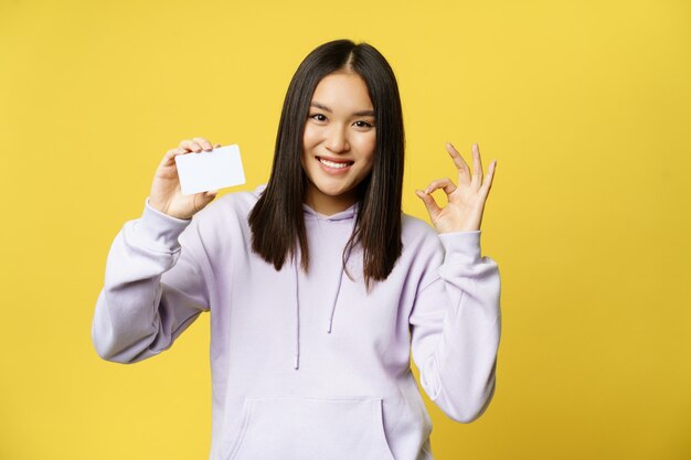 Imagen de niña coreana muestra tarjeta de crédito y signo bien concepto de compra recomendando tienda fondo amarillo ...